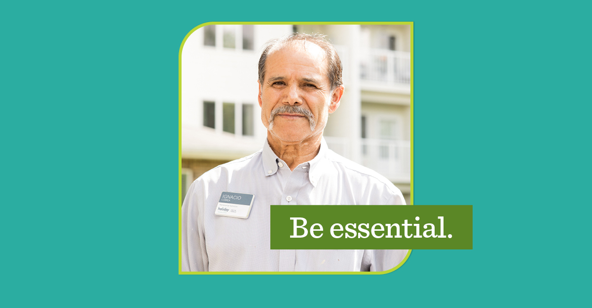 Man with a mustache wearing a name tag and pens in his shirt pocket, standing in front of a building with balconies.